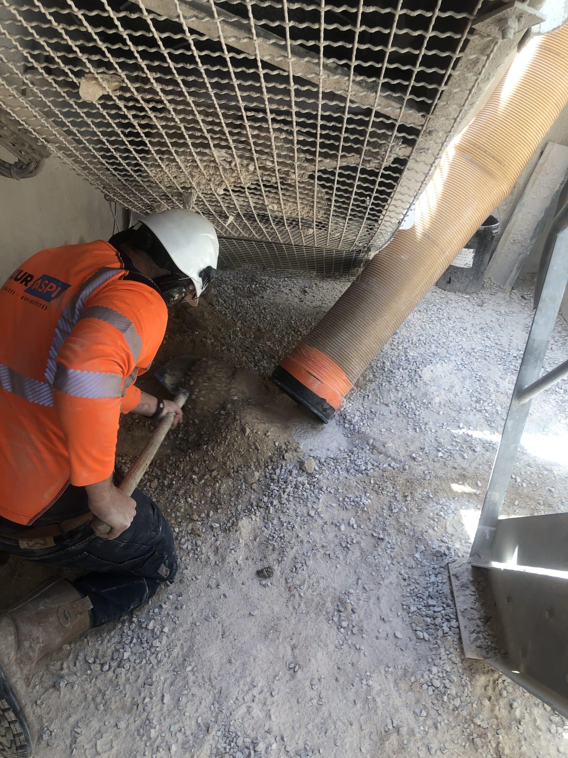 Ouvrier EURASPI avec casque blanc pelletant du gravier vers un tuyau d'aspiration industriel orange en zone de chantier.