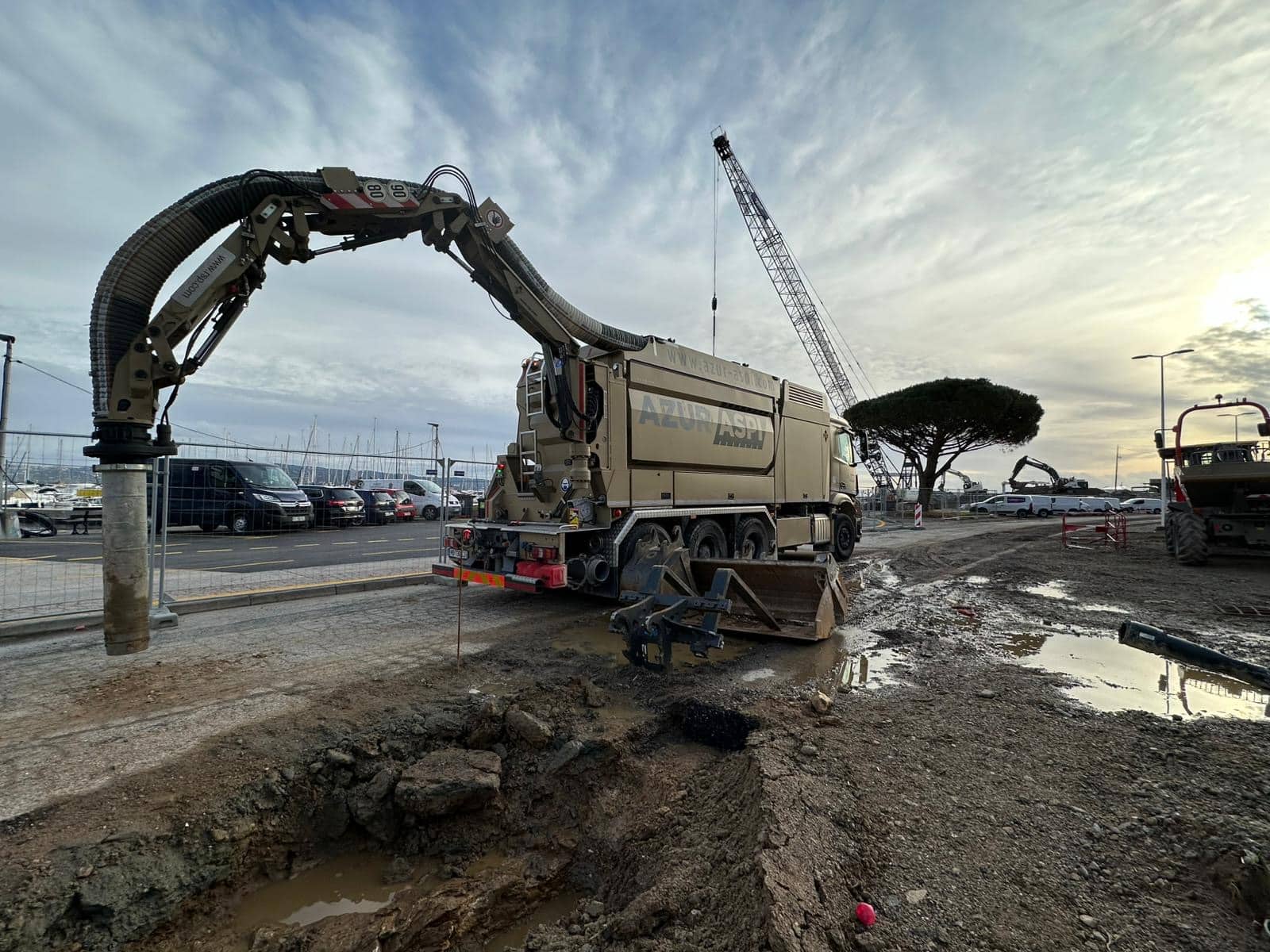 Camion aspirateur AZUR ASPI excavant un terrain boueux sur un chantier à proximité d'un port de plaisance.