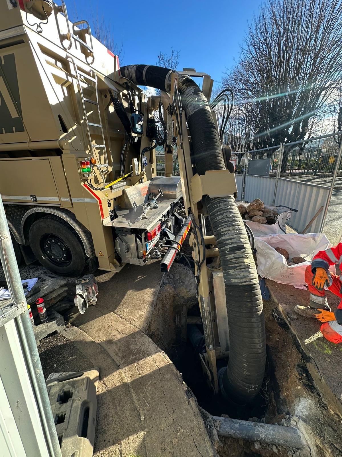Camion aspirateur en opération d'excavation sur un chantier urbain, tuyau dans une tranchée et ouvrier en tenue de sécurité.