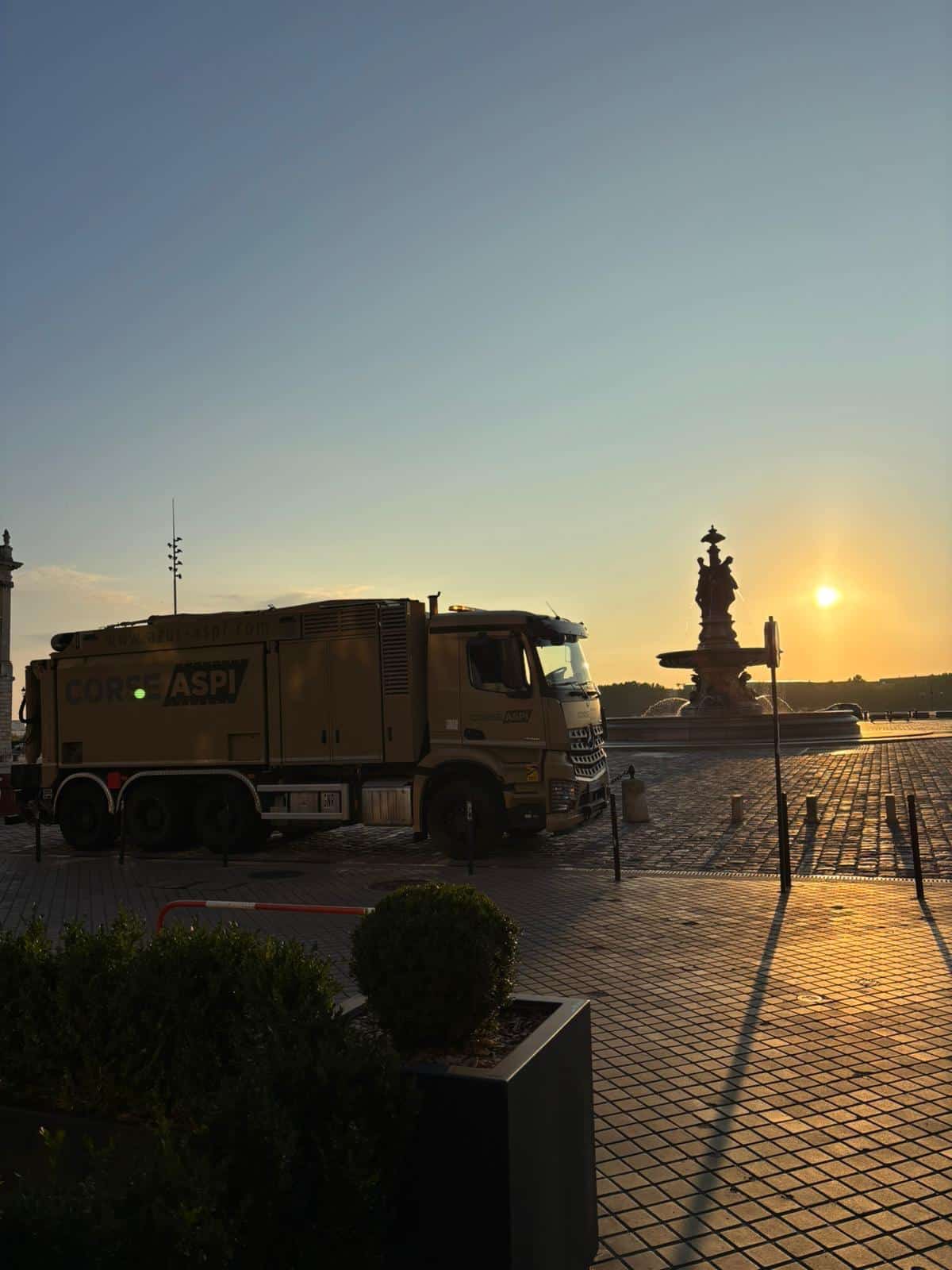 Camion CORSE ASPI devant la fontaine des Trois Grâces à Bordeaux sous un lever de soleil doré.