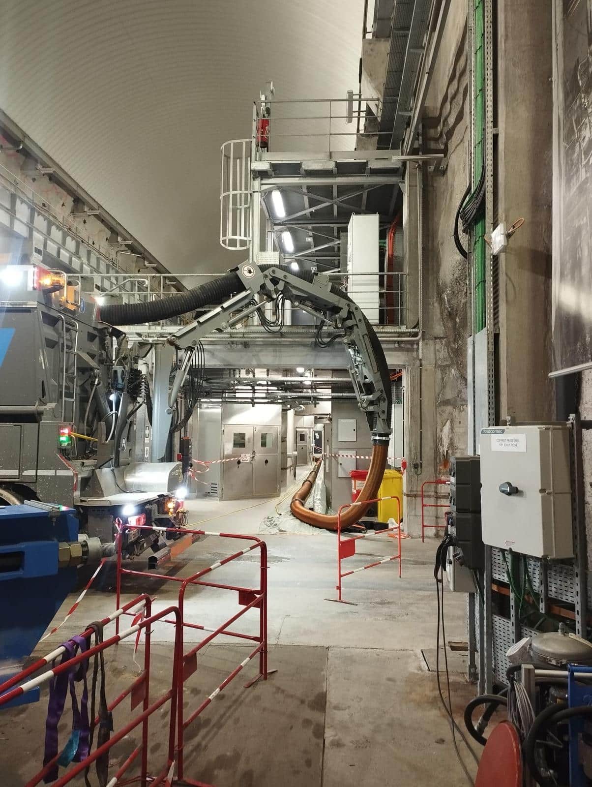 Camion de pompage avec bras articulé et tuyau orange dans un tunnel technique avec boîtiers électriques Socomec.