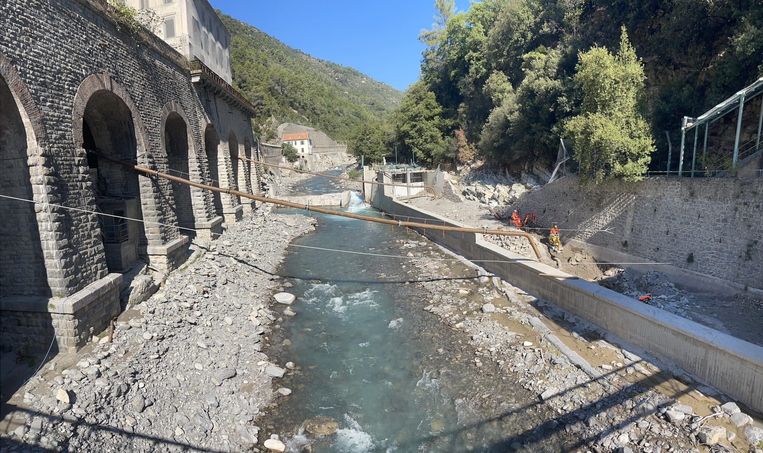 Travaux de maintenance hydraulique sur une rivière de montagne avec canalisation et mur en pierre de taille.