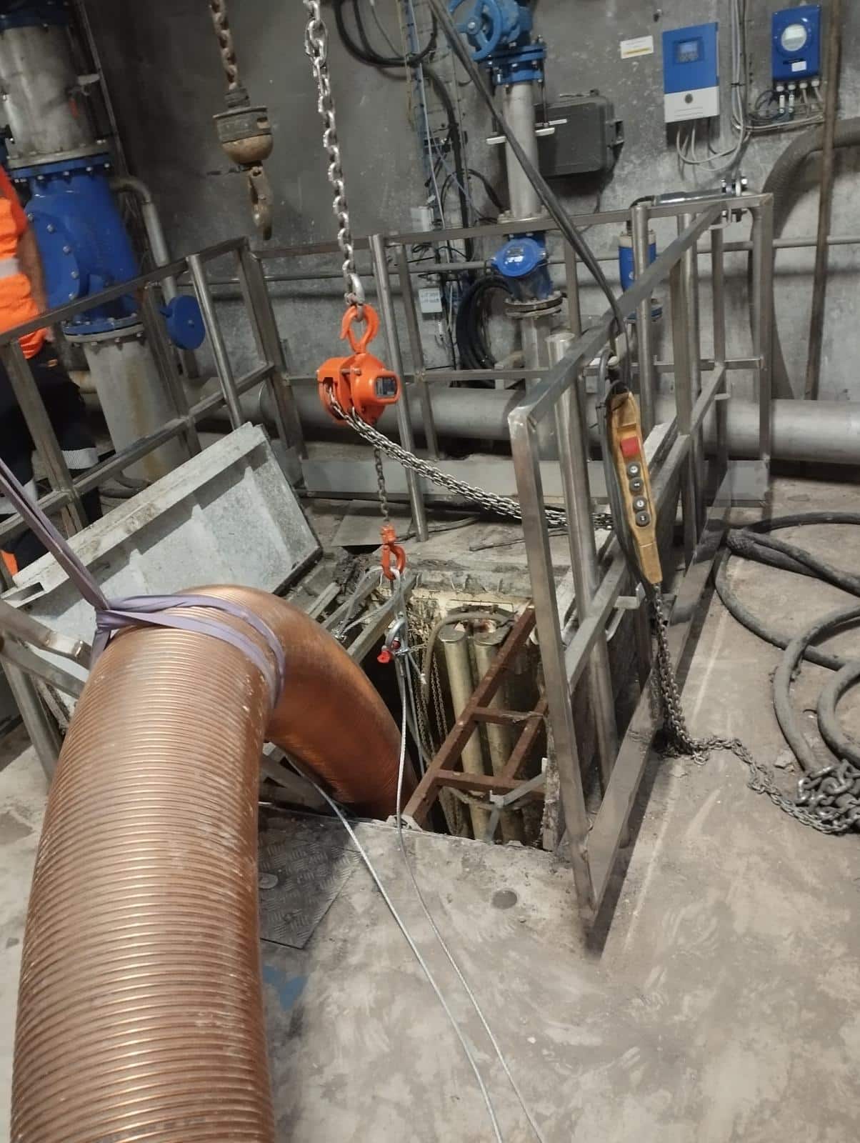Conduit flexible cuivré et palan à chaîne au-dessus d'une trappe ouverte dans un local technique industriel.
