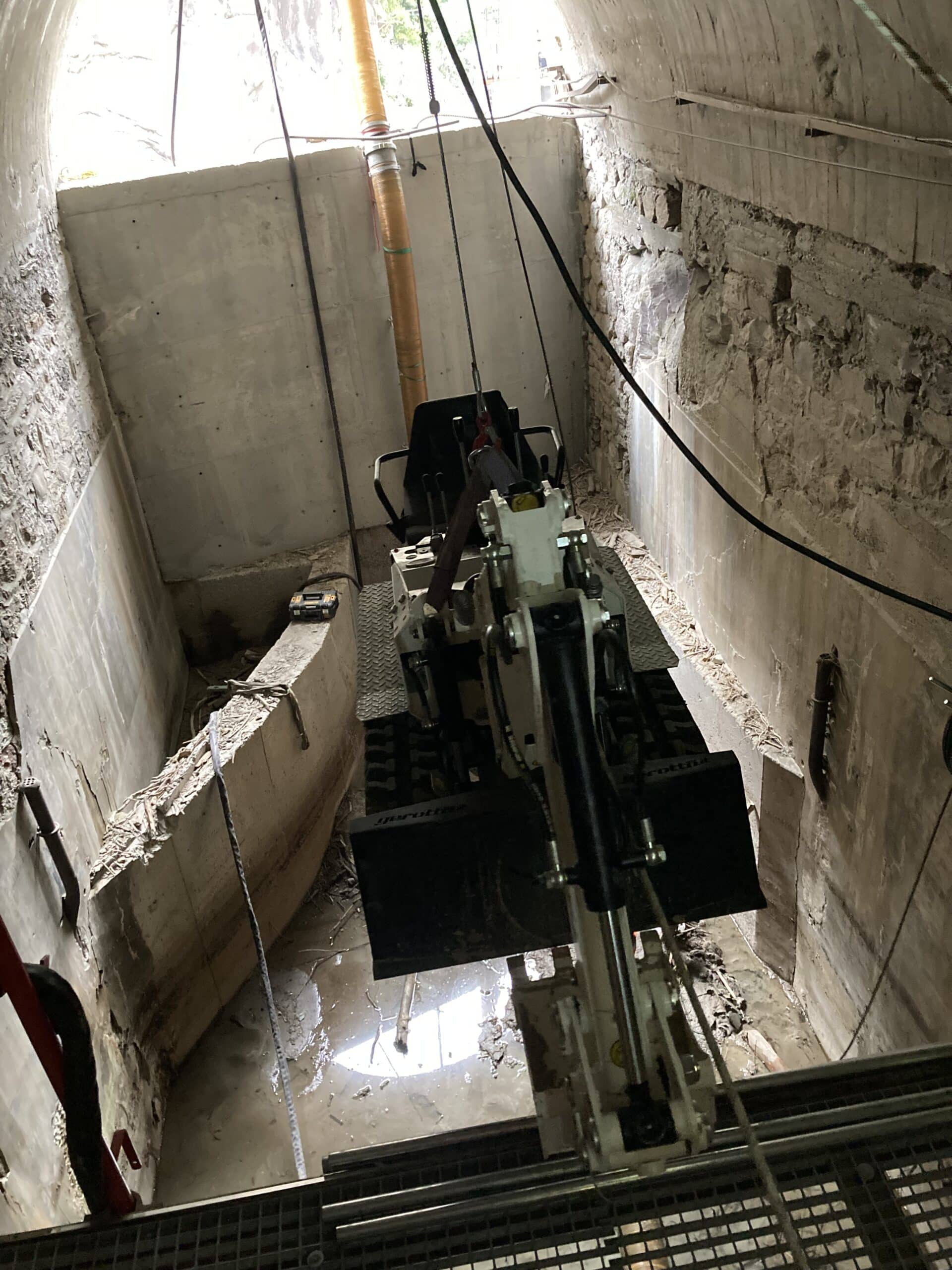 Mini-pelle suspendue par câbles dans un puits de chantier étroit avec parois en béton et sol boueux.