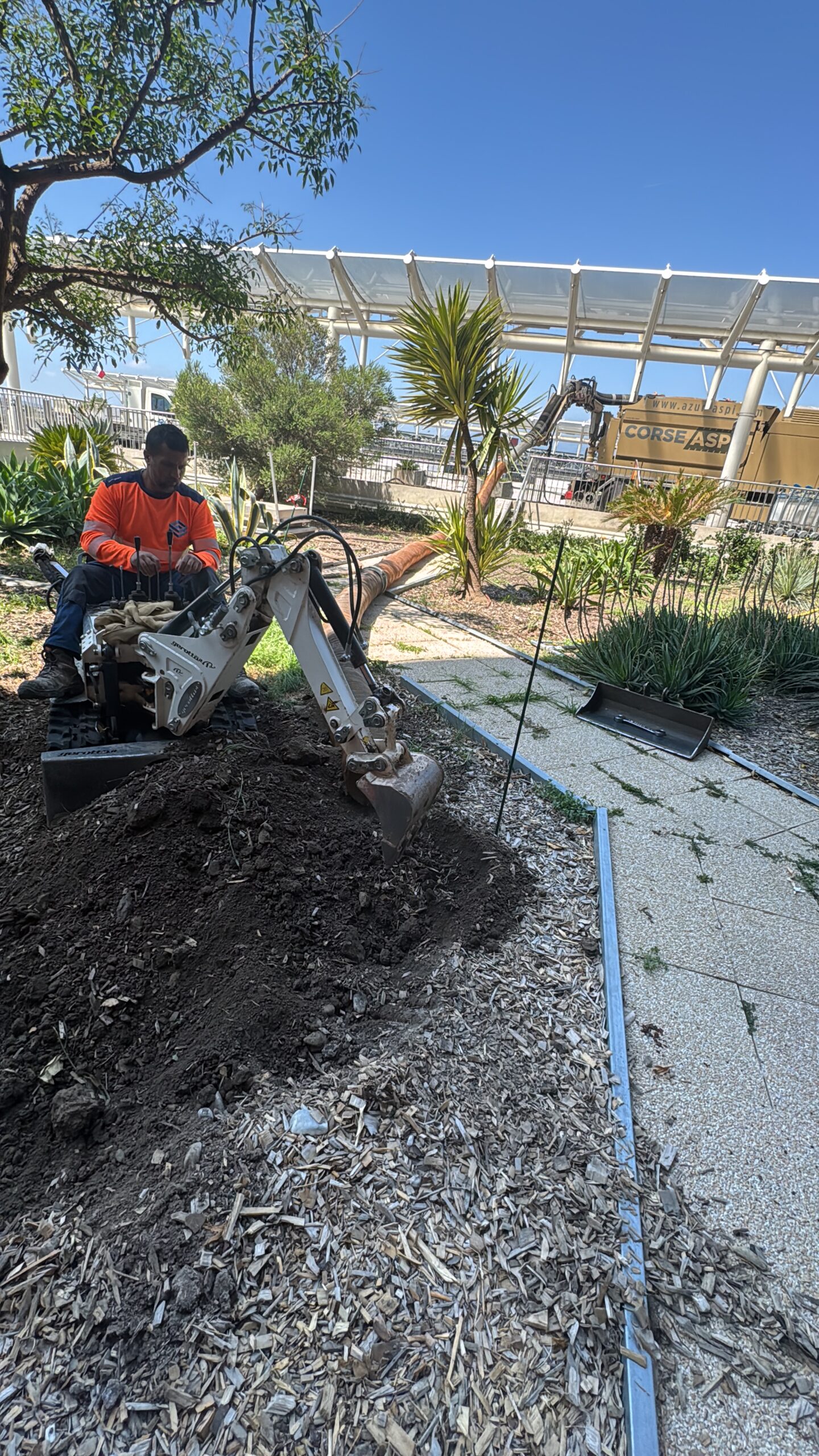 Opérateur maniant une mini-pelle blanche pour creuser un jardin avec camion Corse Asp en arrière-plan.