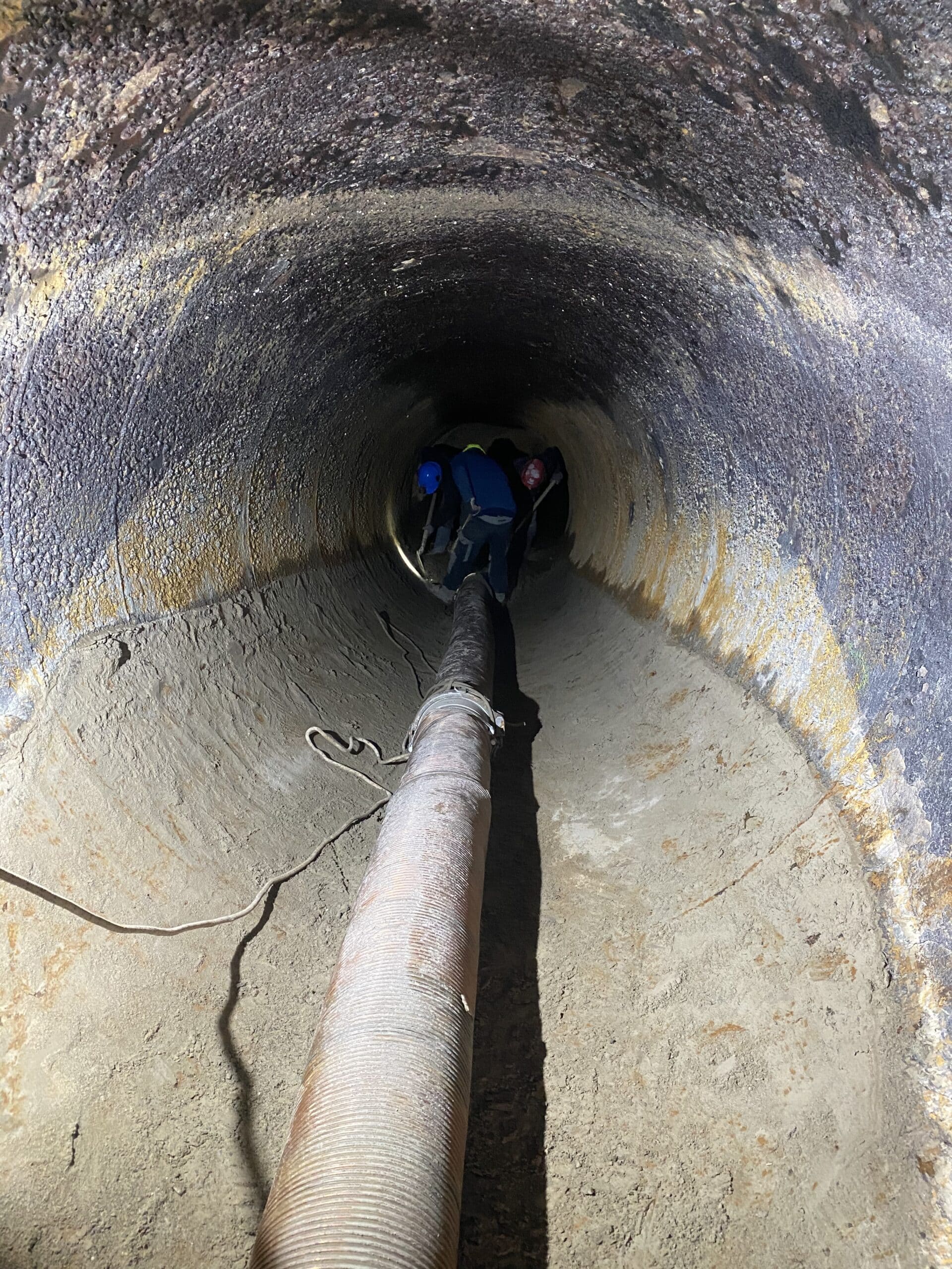 Ouvriers nettoyant une large canalisation souterraine avec un long tuyau d'aspiration flexible.