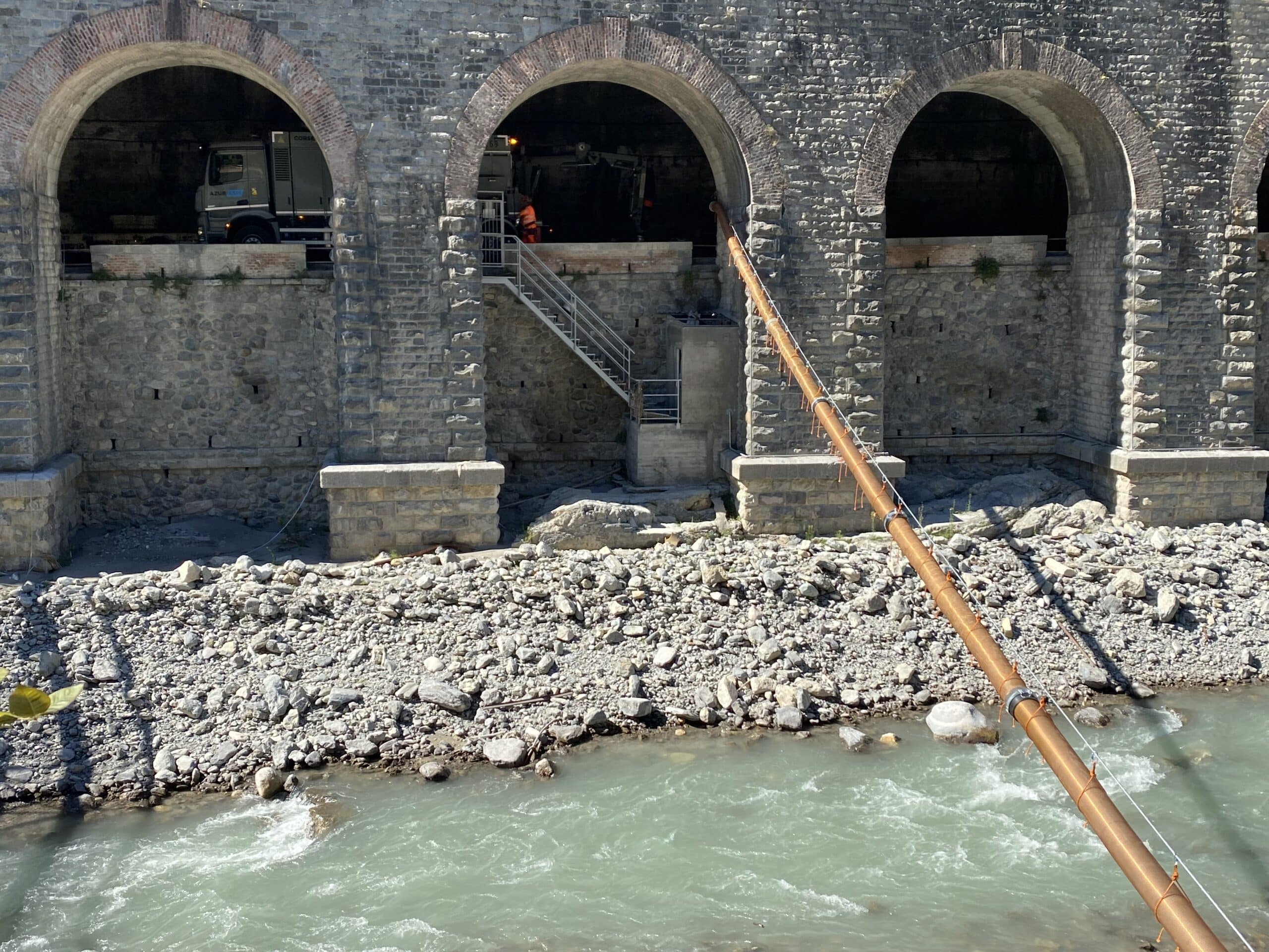 Camion Azur Corse sous les arches d'un pont en pierre enjambant une rivière avec un grand tuyau orange.