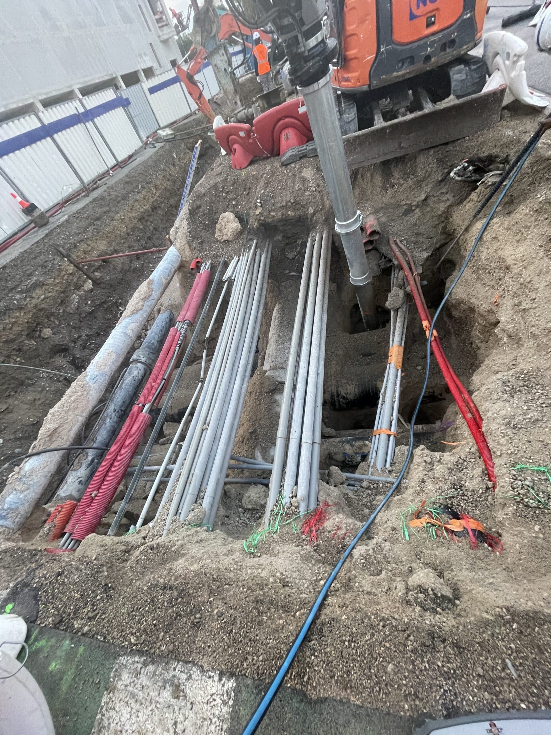 Tranchée de chantier avec canalisations, câbles rouges et conduits gris sous une pelleteuse orange.