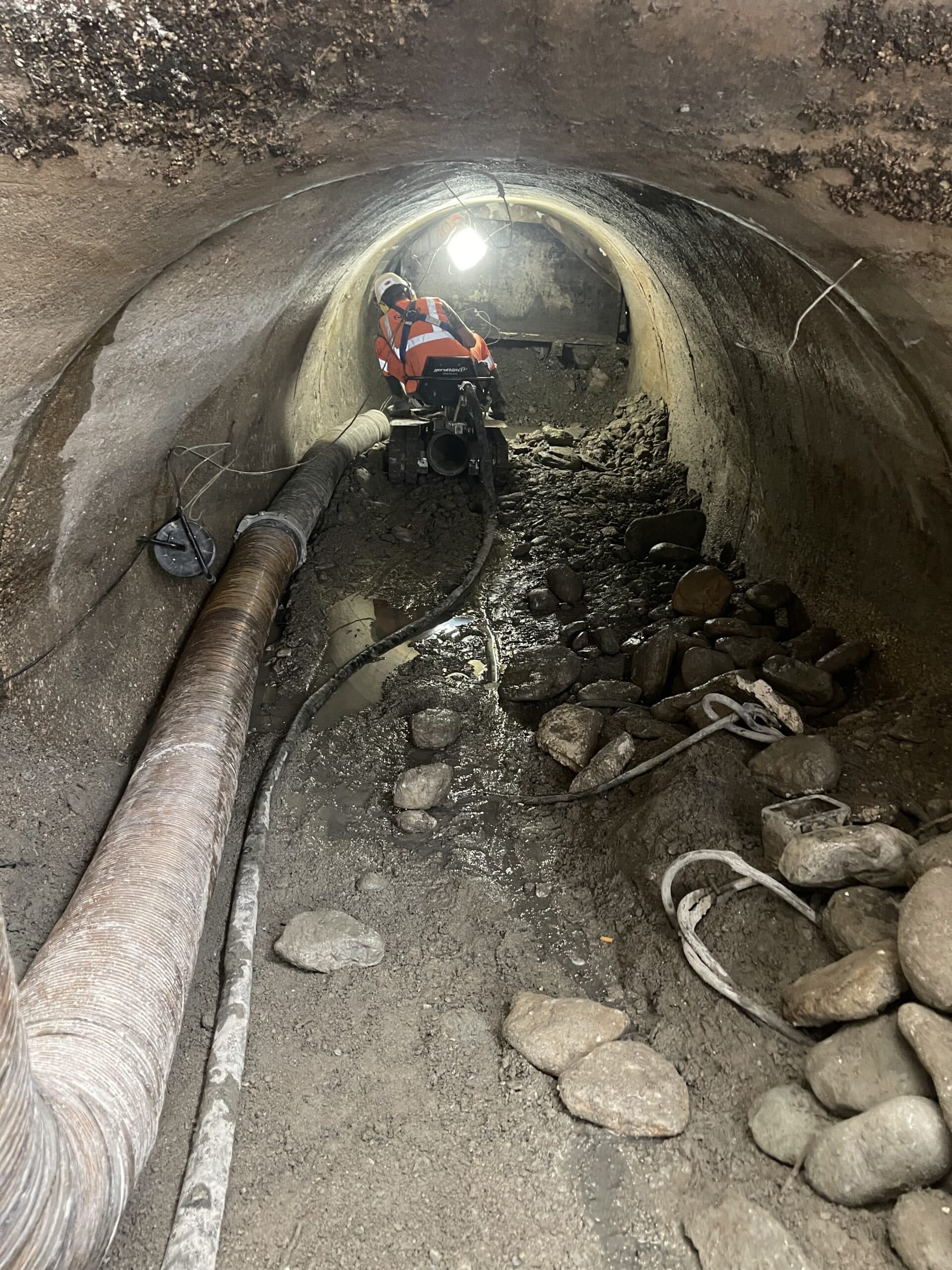 Ouvrier en gilet orange opérant une machine dans un tunnel souterrain avec débris et gros tuyaux de chantier.