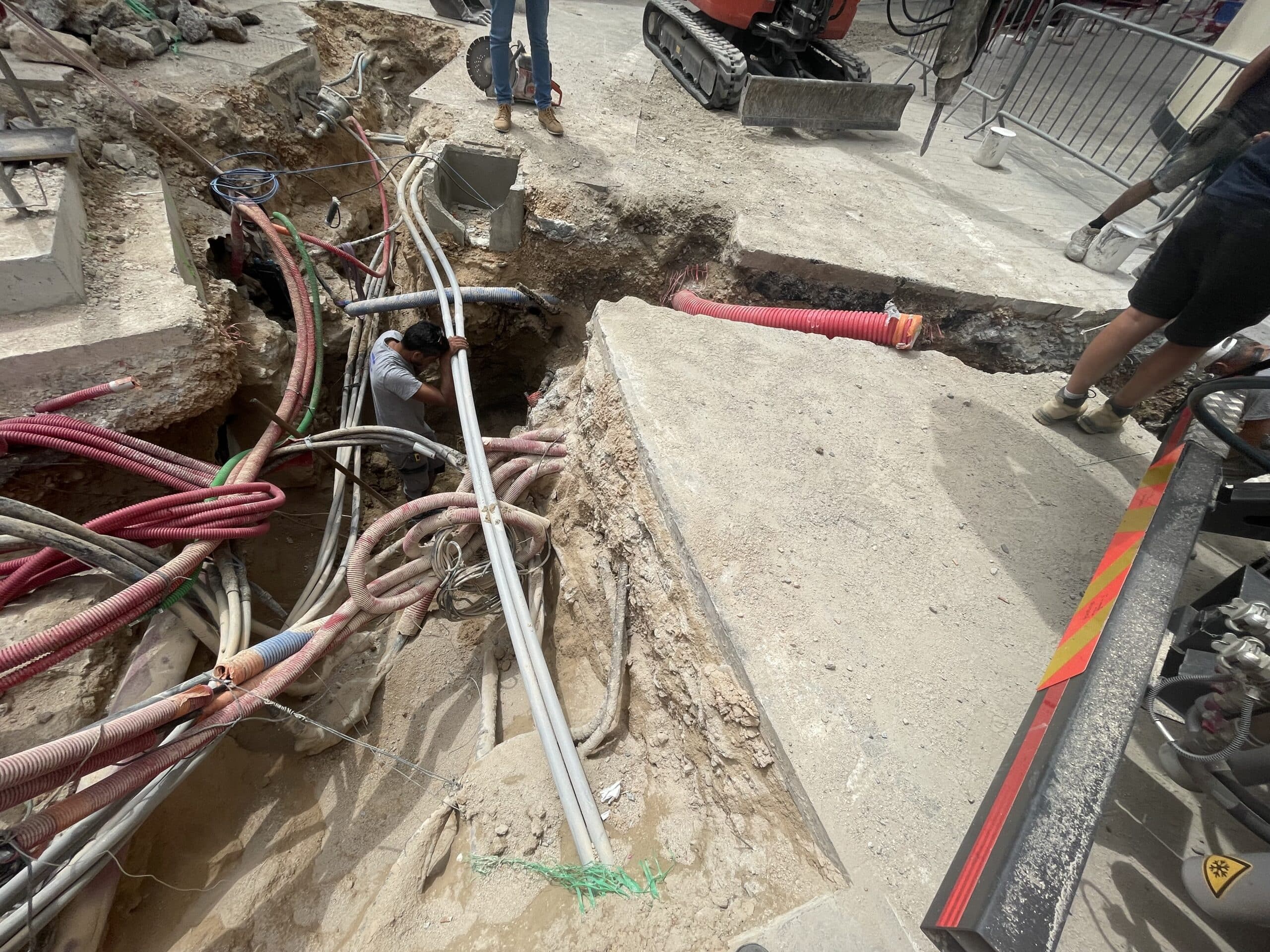 Ouvrier dans une tranchée de chantier avec de nombreux câbles électriques et tuyaux souterrains colorés exposés.