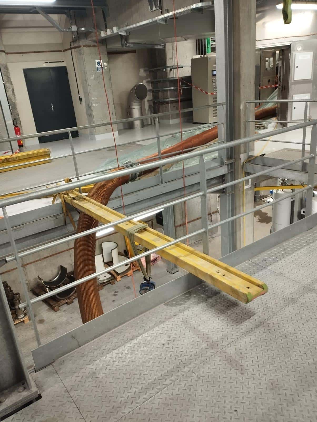 Gros tuyau flexible marron sur une passerelle industrielle, soutenu par une poutre jaune lors d'une maintenance.