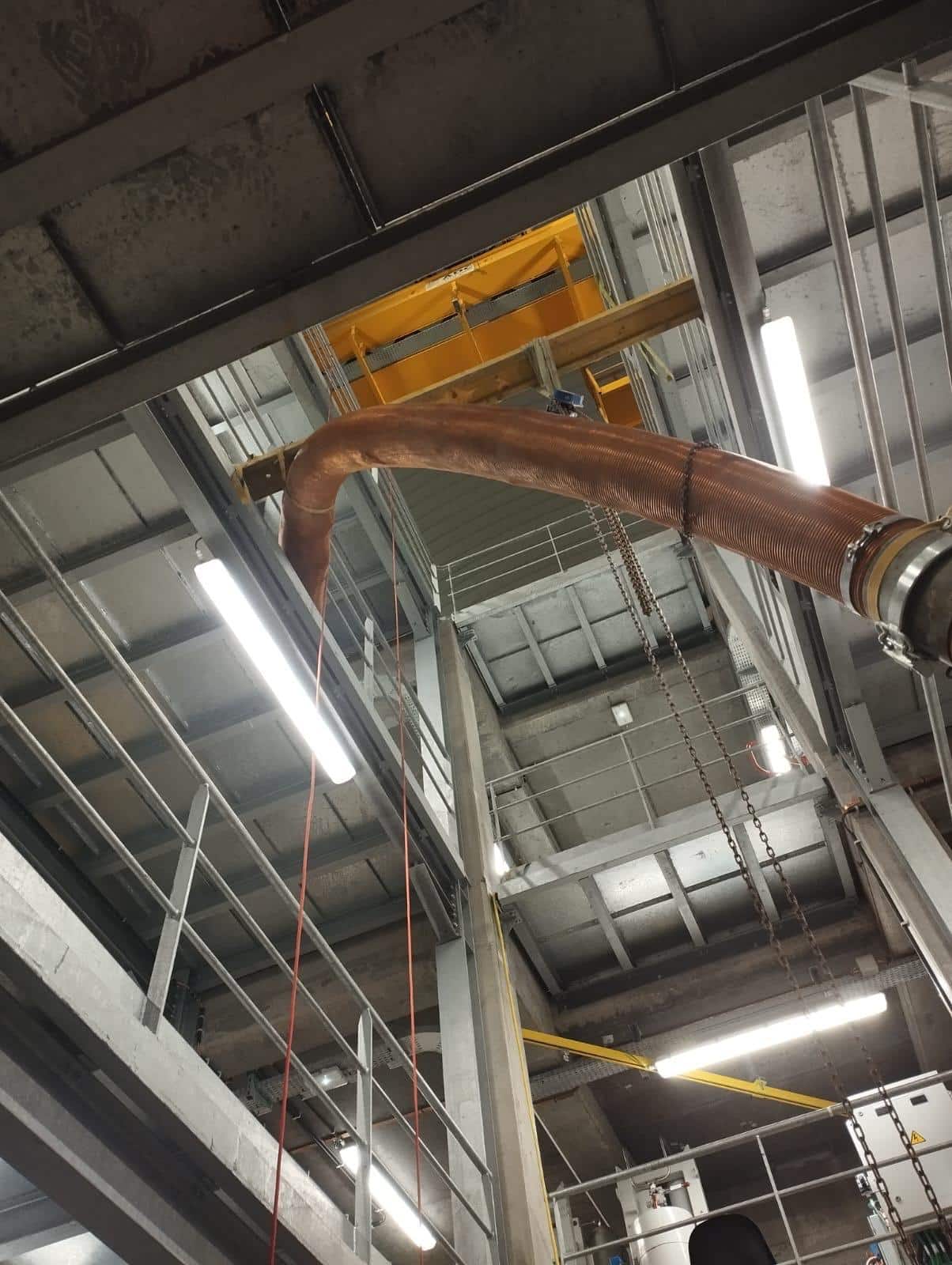 Gros tuyau flexible cuivré traversant une structure industrielle métallique multi-niveaux avec éclairage LED intense.