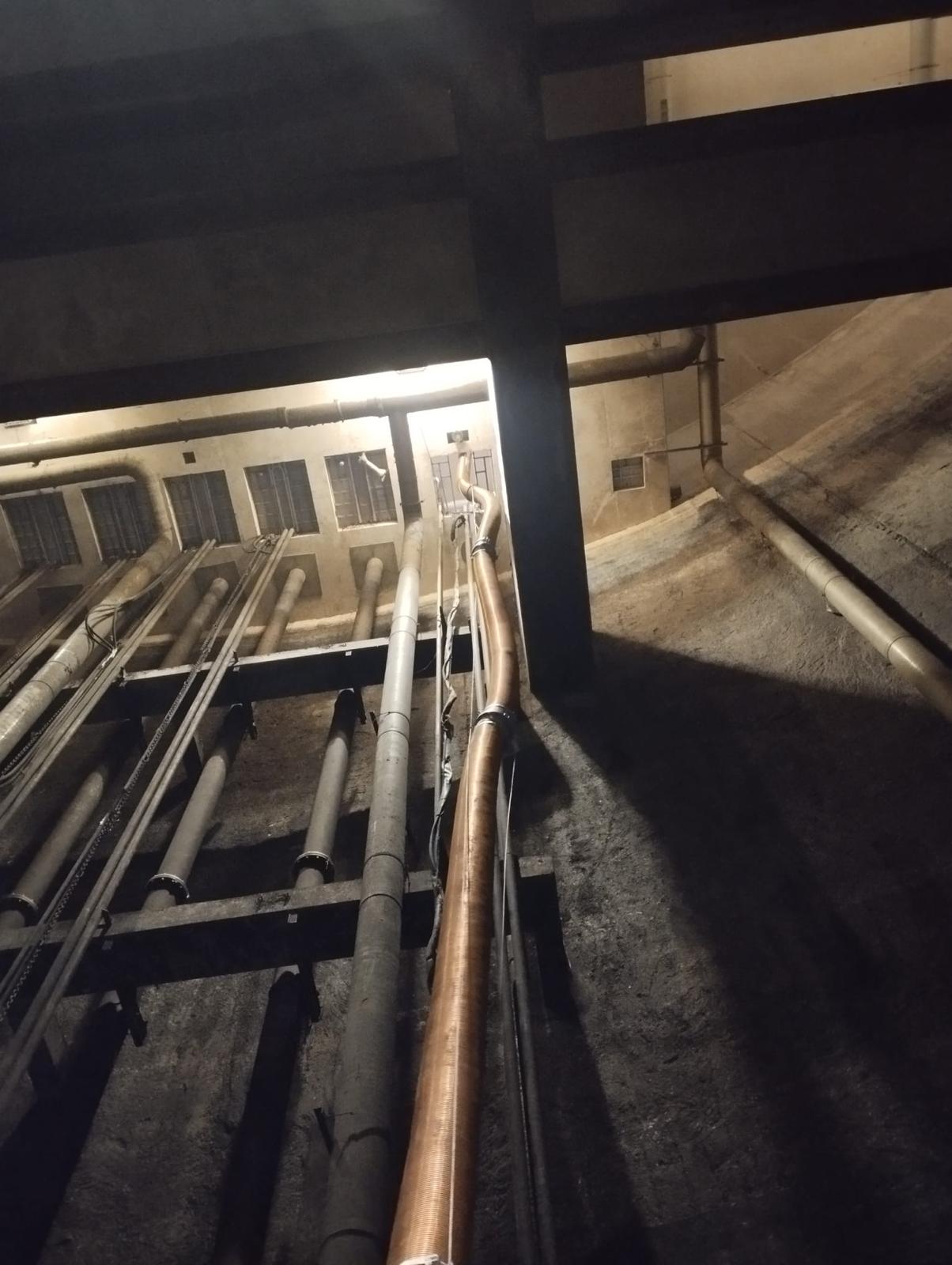 Tuyaux industriels et conduits métalliques dans un espace souterrain sombre avec une structure de support en acier.
